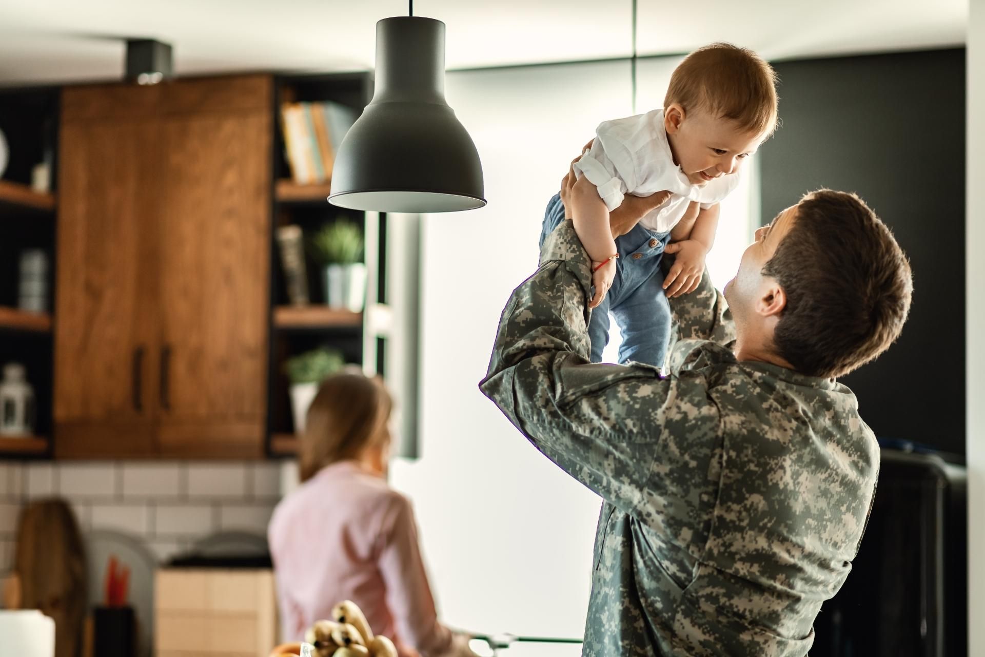 Man lifting a baby indoors while a child stands nearby in a sunlit home kitchen