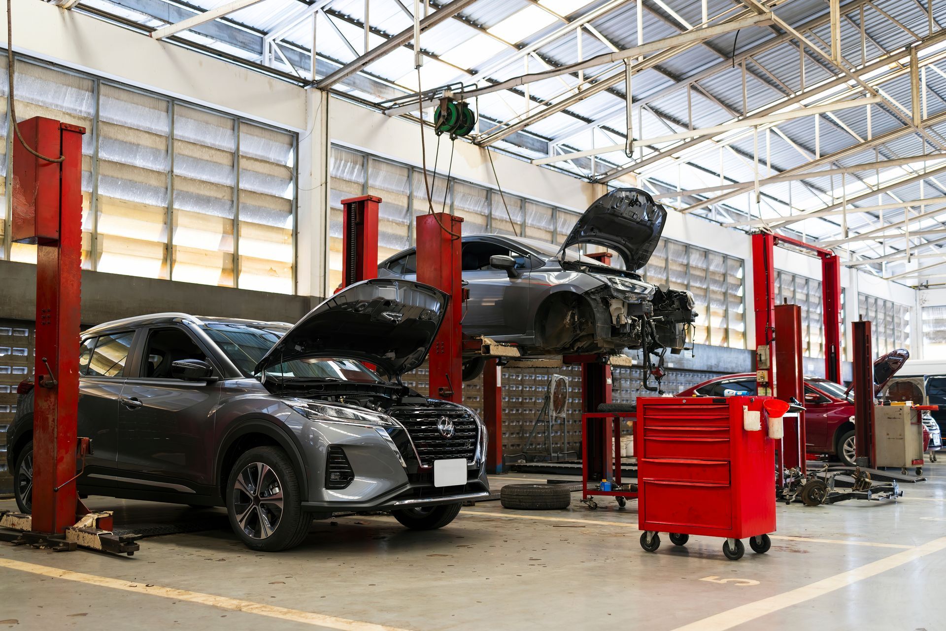 Two SUVs in an auto repair shop with hoods open, red lift posts, and a tool cart.