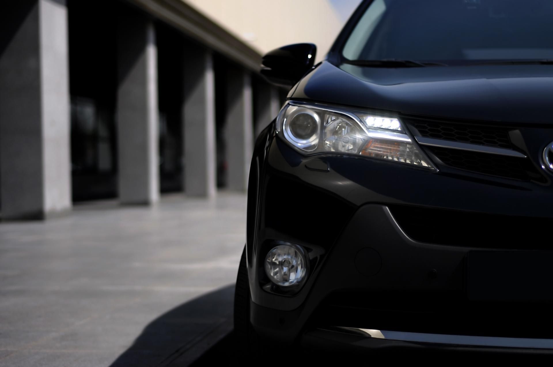 Front of a black car parked beside a concrete building, showing the headlight and grille