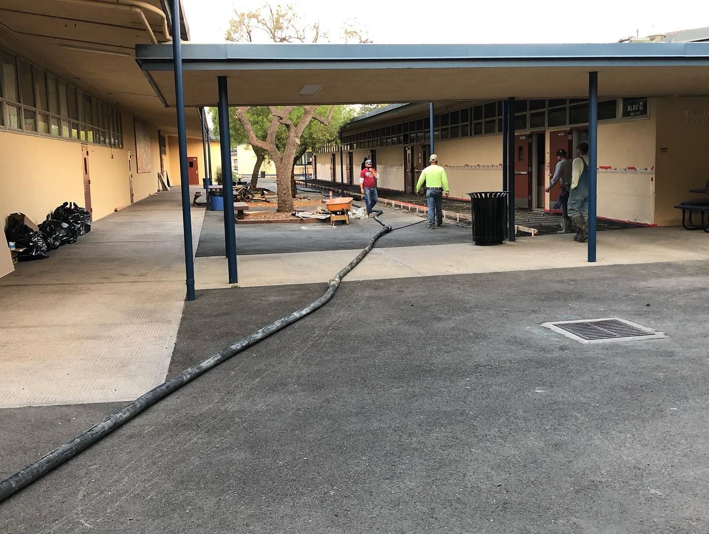 Outdoor walkway between buildings with workers and equipment, one worker in a yellow vest near a hose.