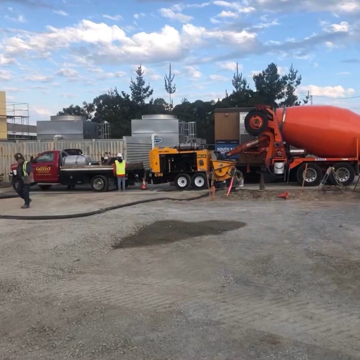 Construction trucks and workers beside a large orange cement mixer on a gravel lot under a cloudy sky