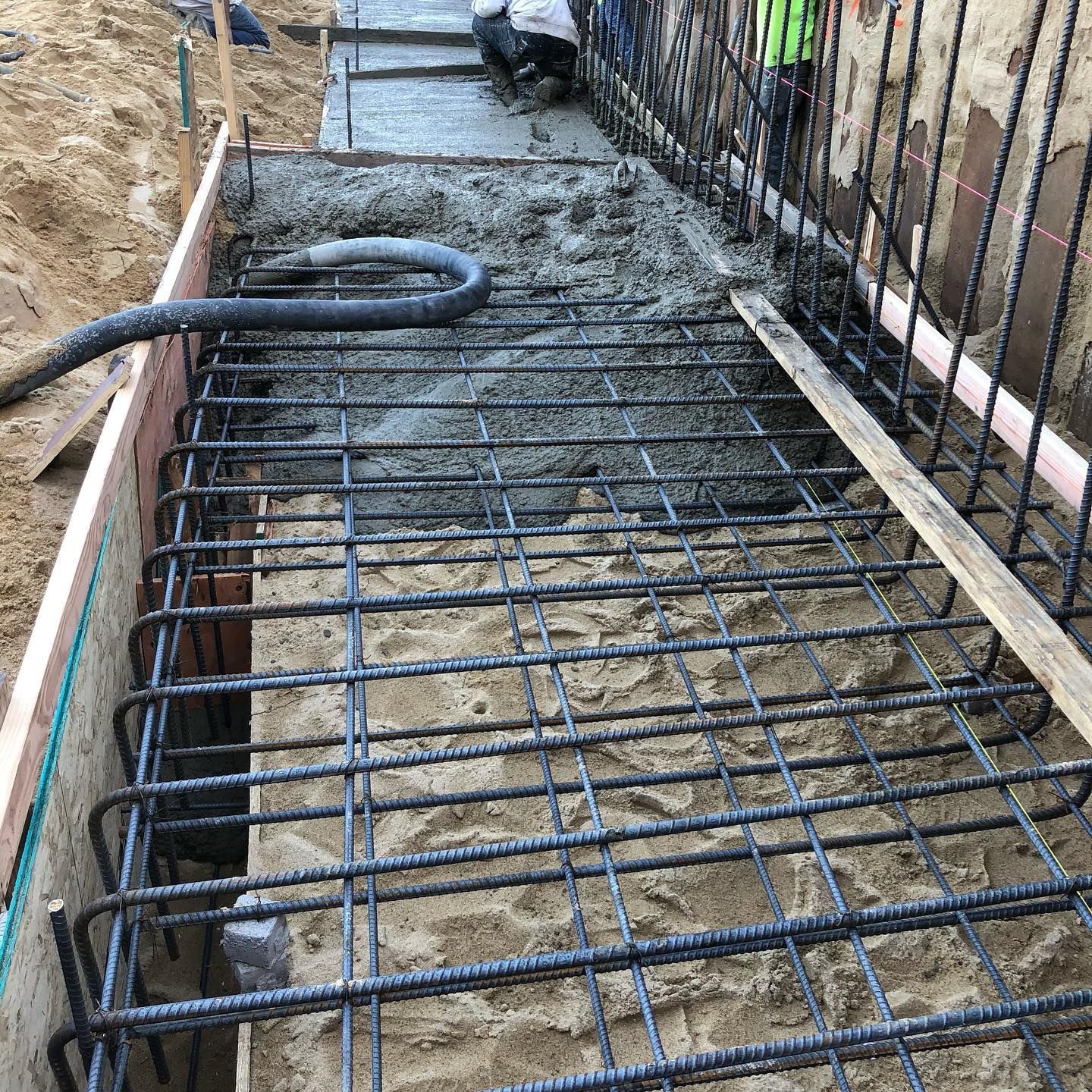 Rebar grid laid over a narrow concrete trench at a construction site, with wooden forms and hoses nearby
