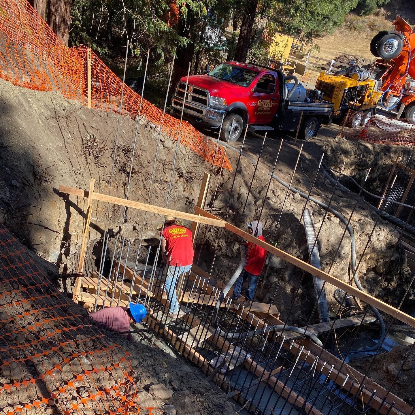 Construction workers in a trench beside a red truck and heavy equipment, with orange safety fencing nearby