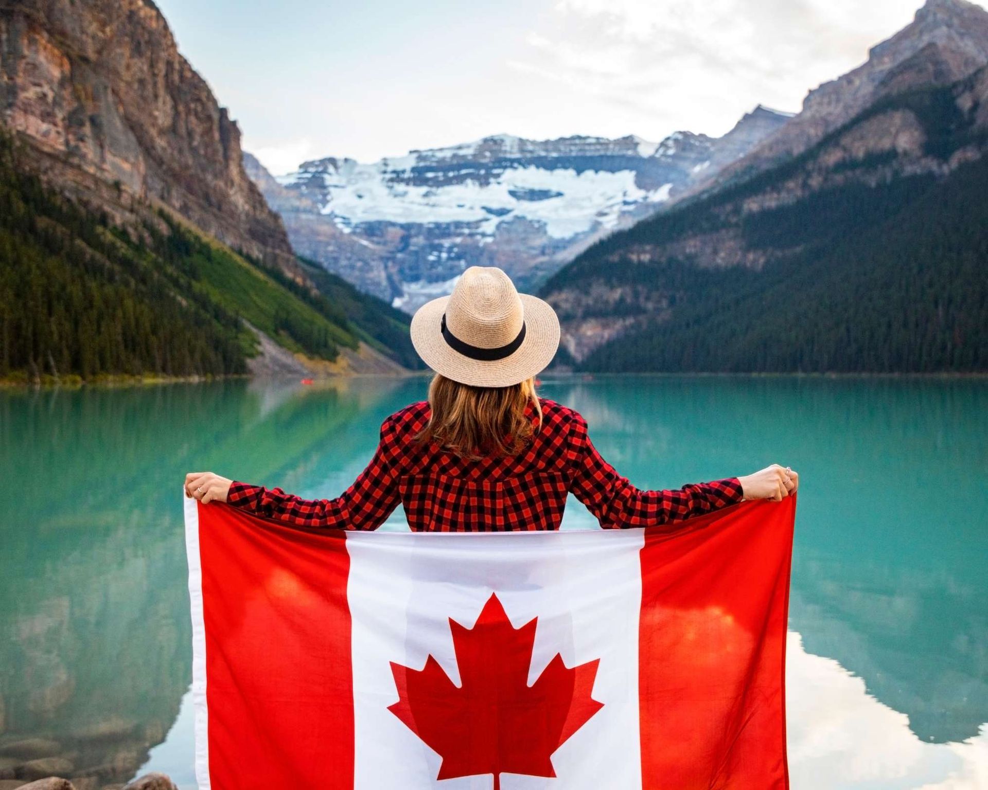 Person holding Canadian flag, facing a turquoise lake, mountains, and snow.