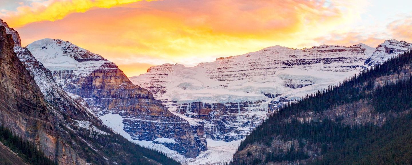 Snow-covered mountains under a vibrant, orange and yellow sunset. Dark green trees frame the scene.