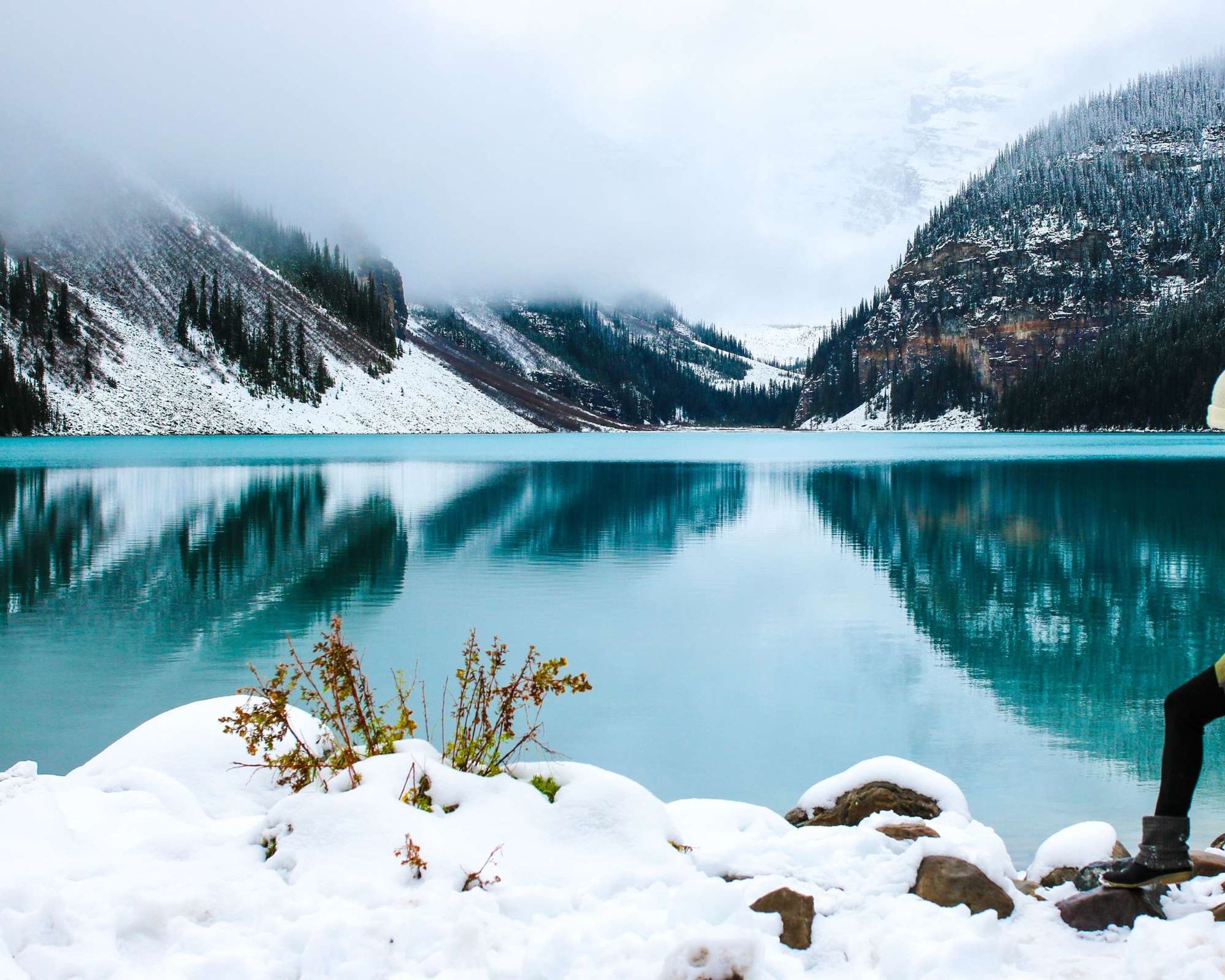 Snowy lake with turquoise water reflects mountains under overcast sky.