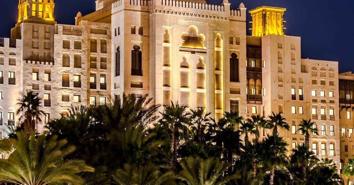 Lit-up, ornate white building with architectural details, palm trees in front, at night.