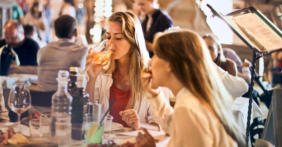 Two women dining at a restaurant, one drinking, the other eating; blurred background of diners.