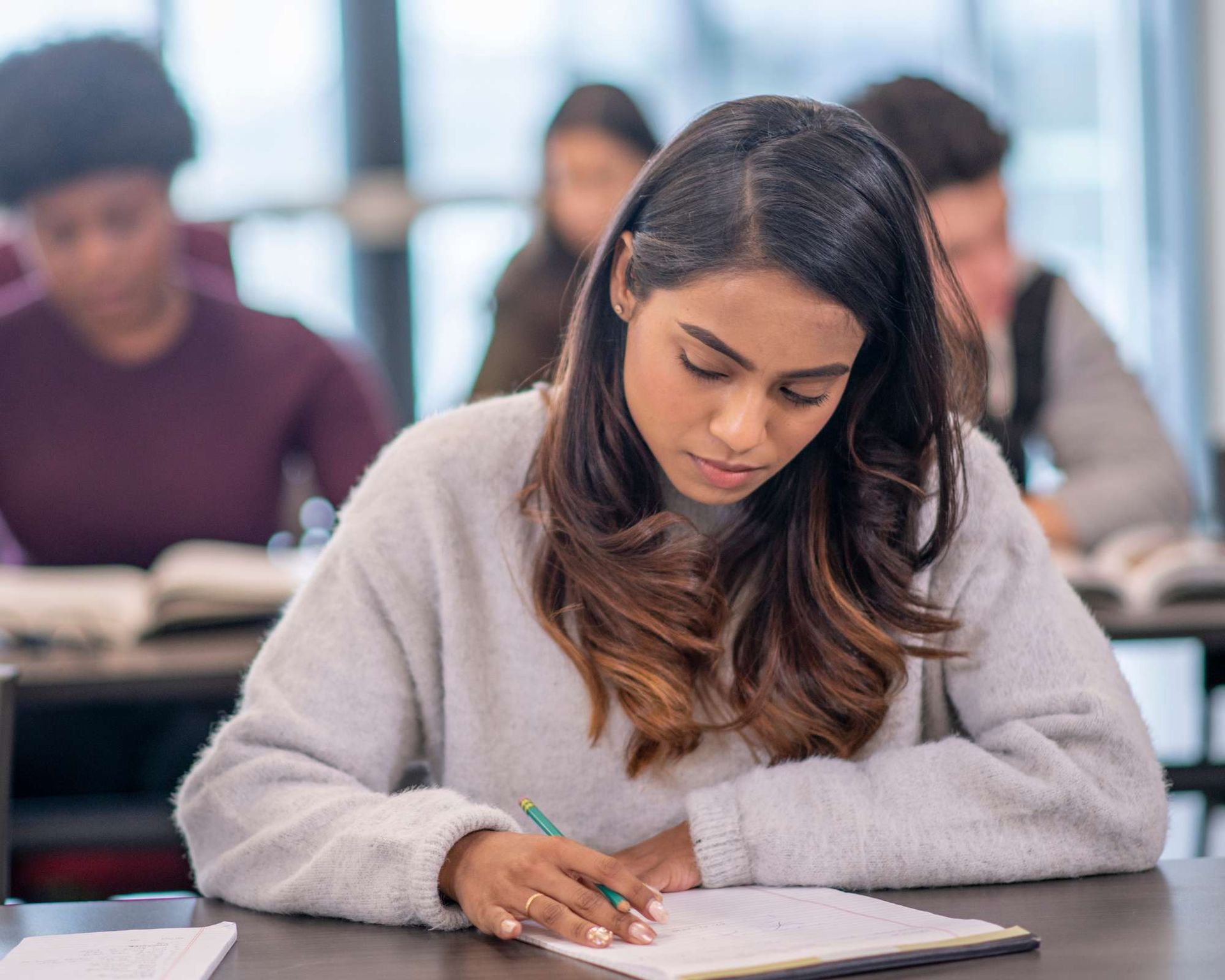 Student writing in notebook at desk in classroom, other students blurred in background.
