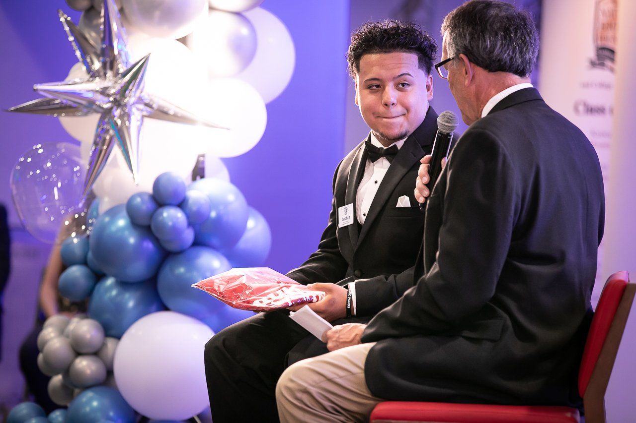 Two men seated, one in a tuxedo holding a gift, the other with a microphone. Balloons in background.