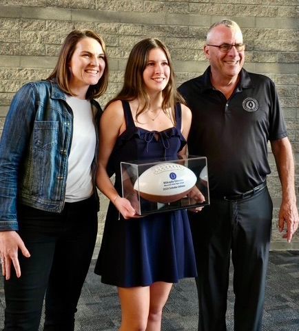 Woman, girl, and man smiling, holding football award in front of a wall.