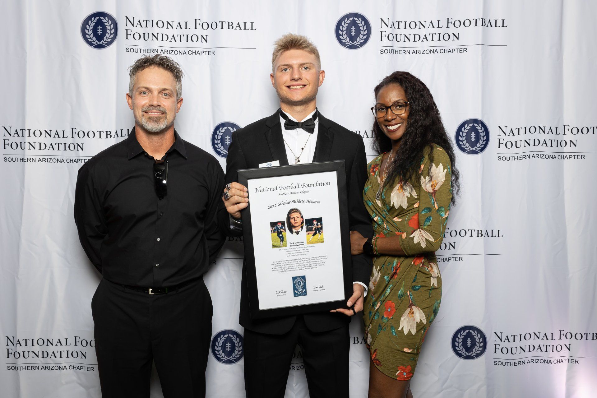 Three people pose with a framed document. Man in a tux holds it. Two others flank him. National Football Foundation backdrop.