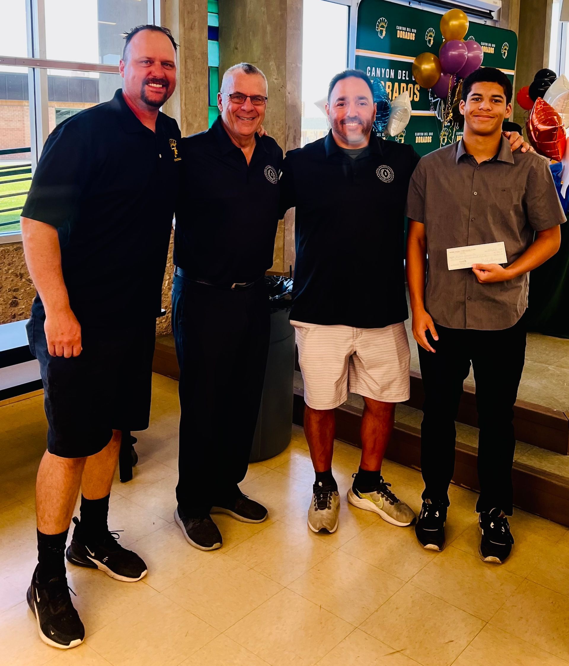 Four men pose indoors, one holding a check.  They're smiling, near decorations and a building exterior.