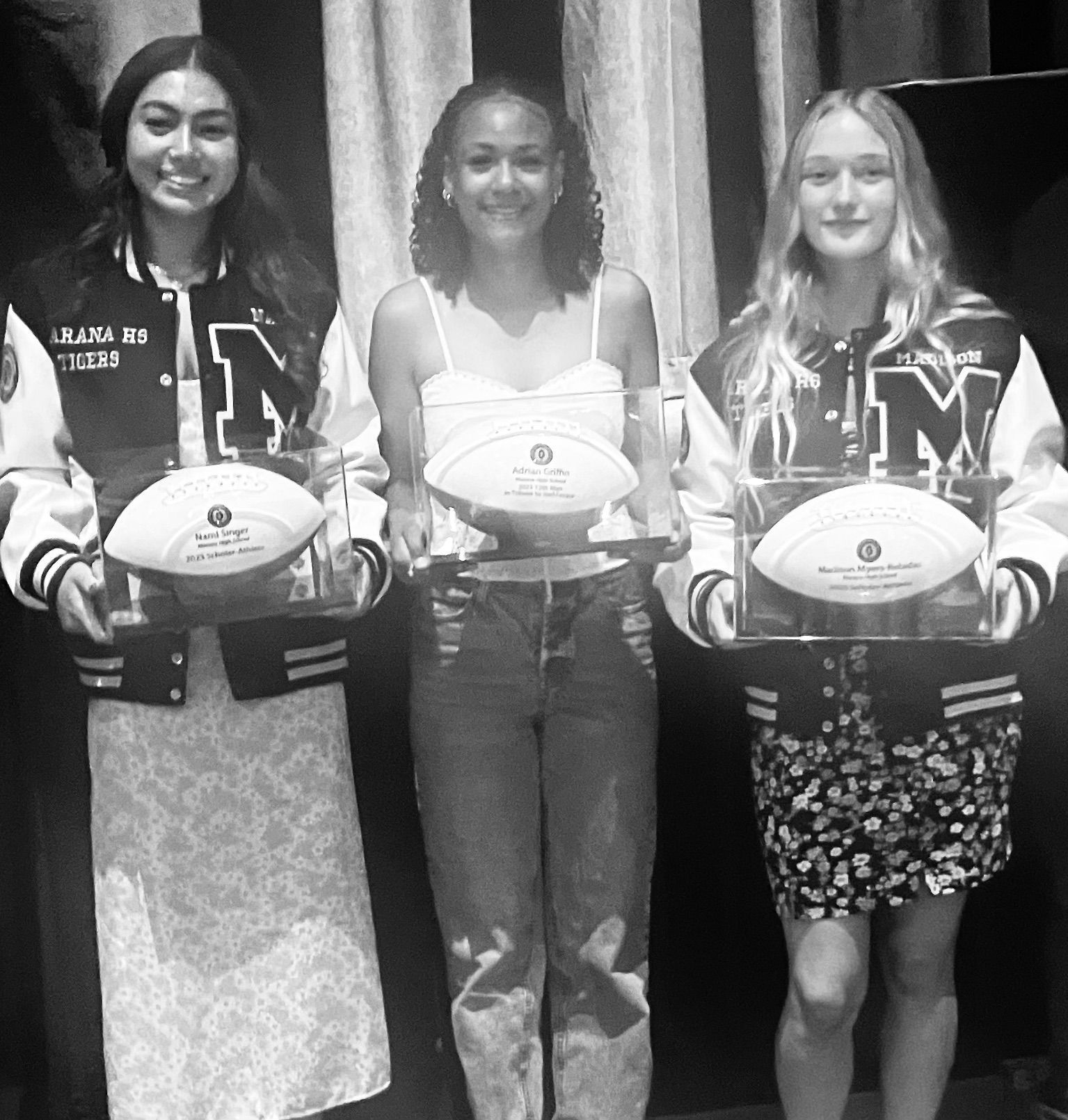 Three people holding trophies, wearing letterman jackets and smiling, standing on a stage.