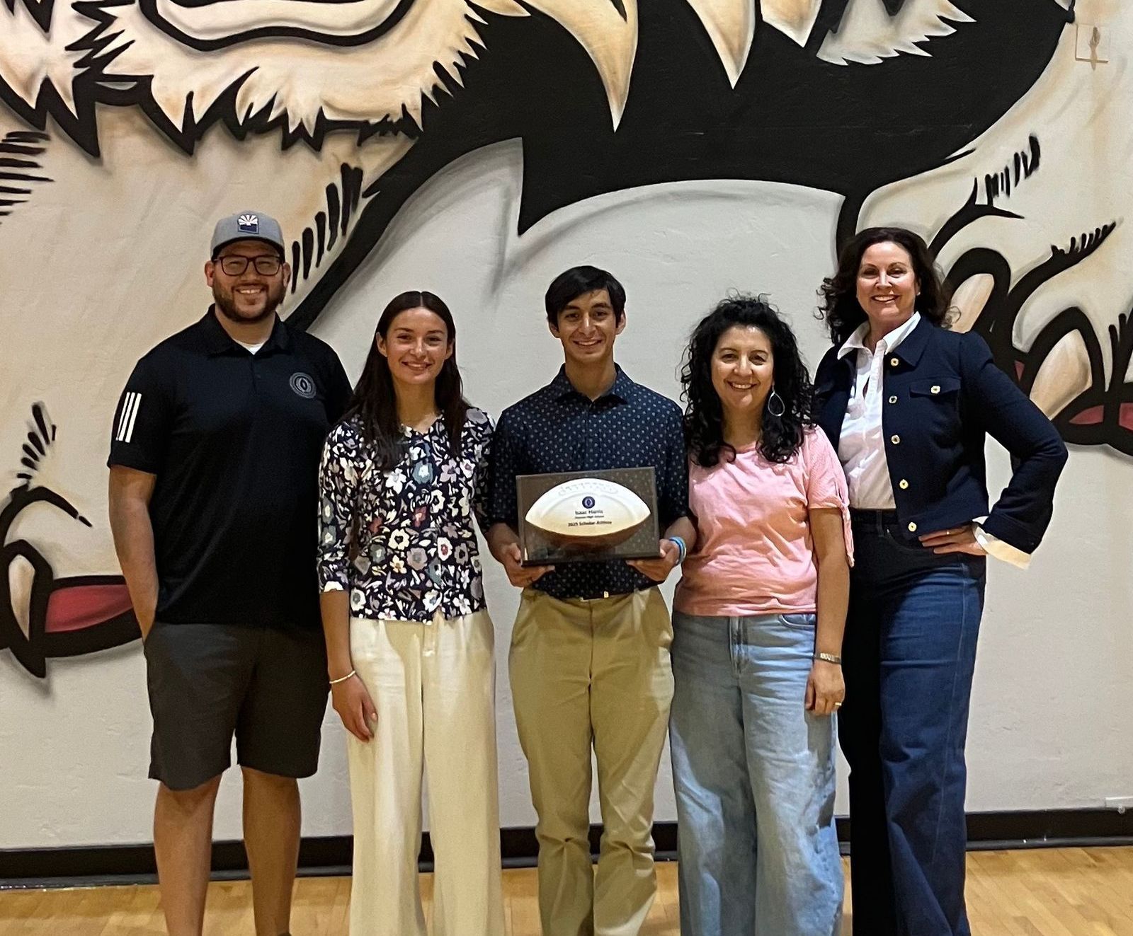 Five people posing in front of a school mural. A person holds an award shaped like a football.