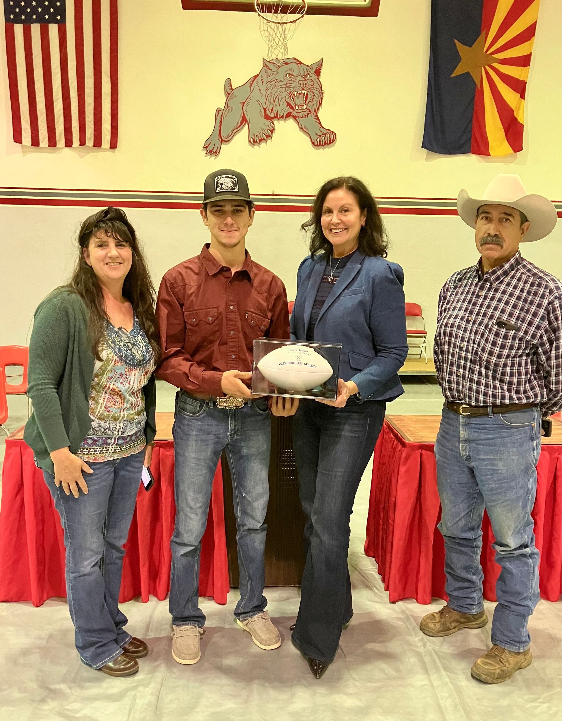 Four people standing, holding a football. Two women and two men pose under flags and a school symbol.