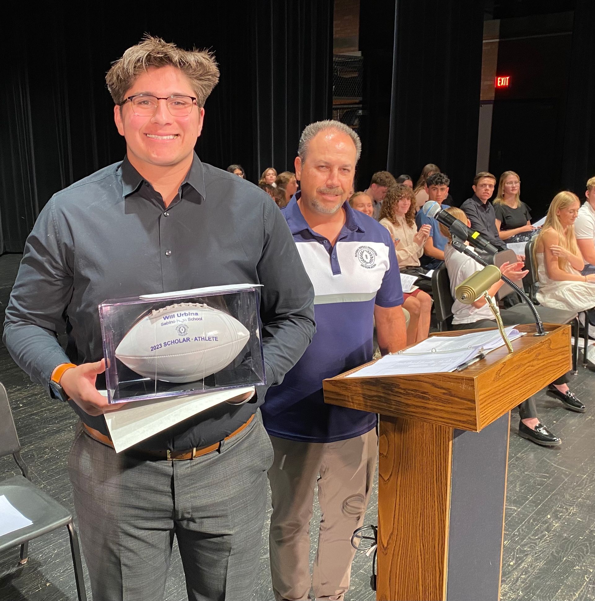 Man holding a trophy football next to a man at a podium with audience in a theater.