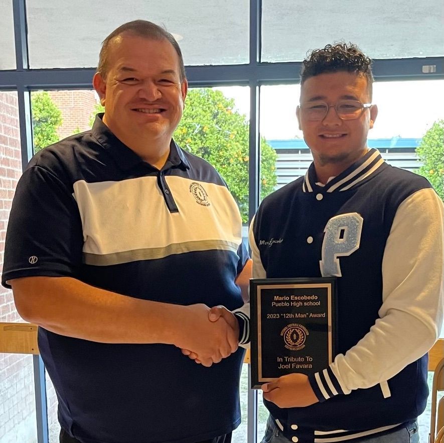 Man hands award to a young person. Both smiling, near a window. The award is a plaque.