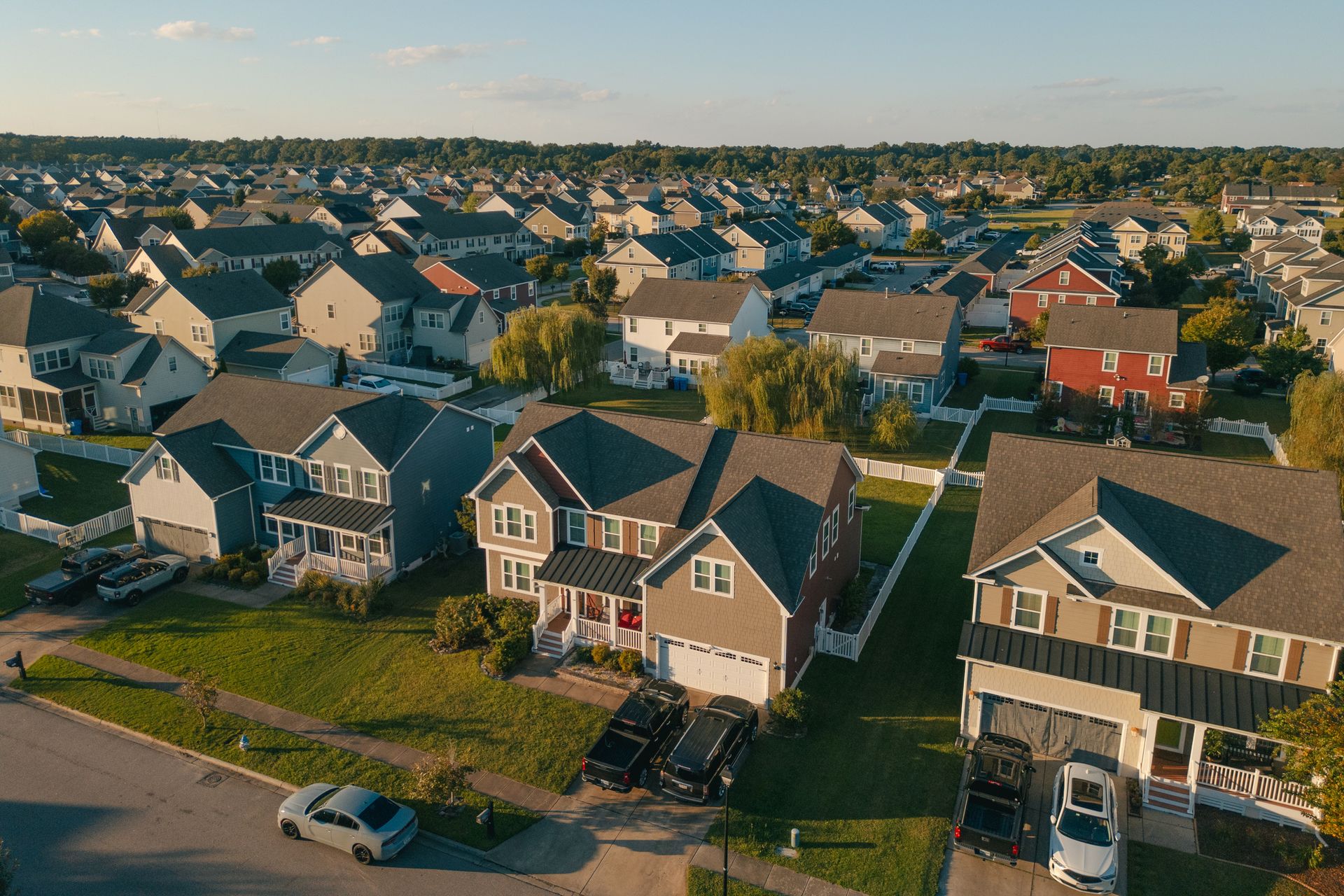 Aerial view of a suburban neighborhood with rows of houses, green lawns, and cars parked in driveways under a blue sky.
