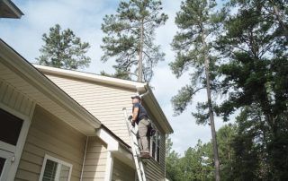Person on a ladder cleaning gutters on a light-colored house, with tall trees in the background.
