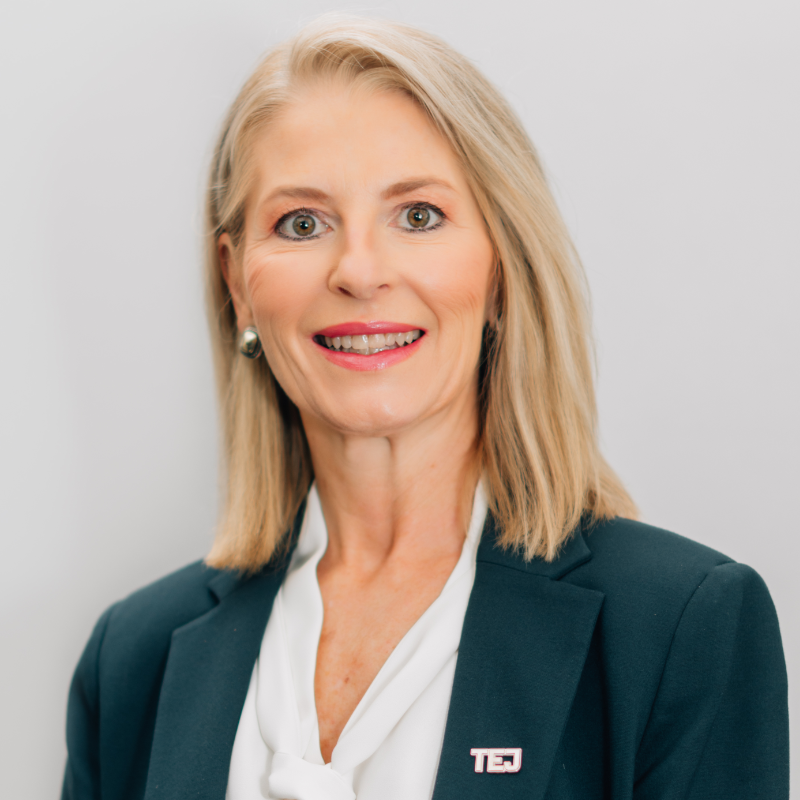 Headshot of a blonde woman in a teal blazer and white blouse, smiling against a light background