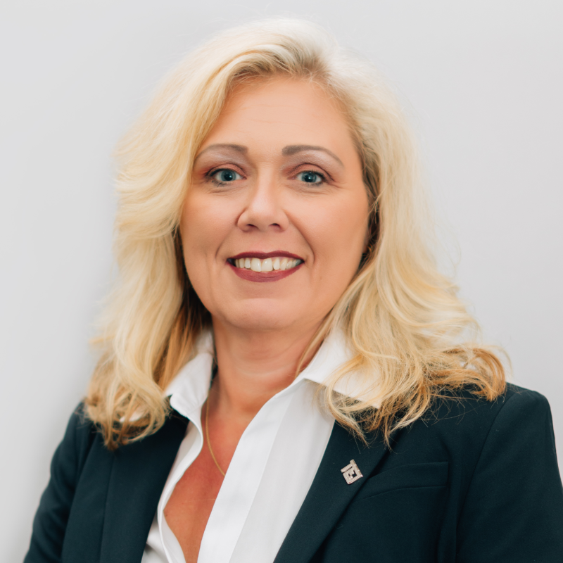 Professional headshot of a smiling blonde woman in a dark blazer and white blouse on a light gray background