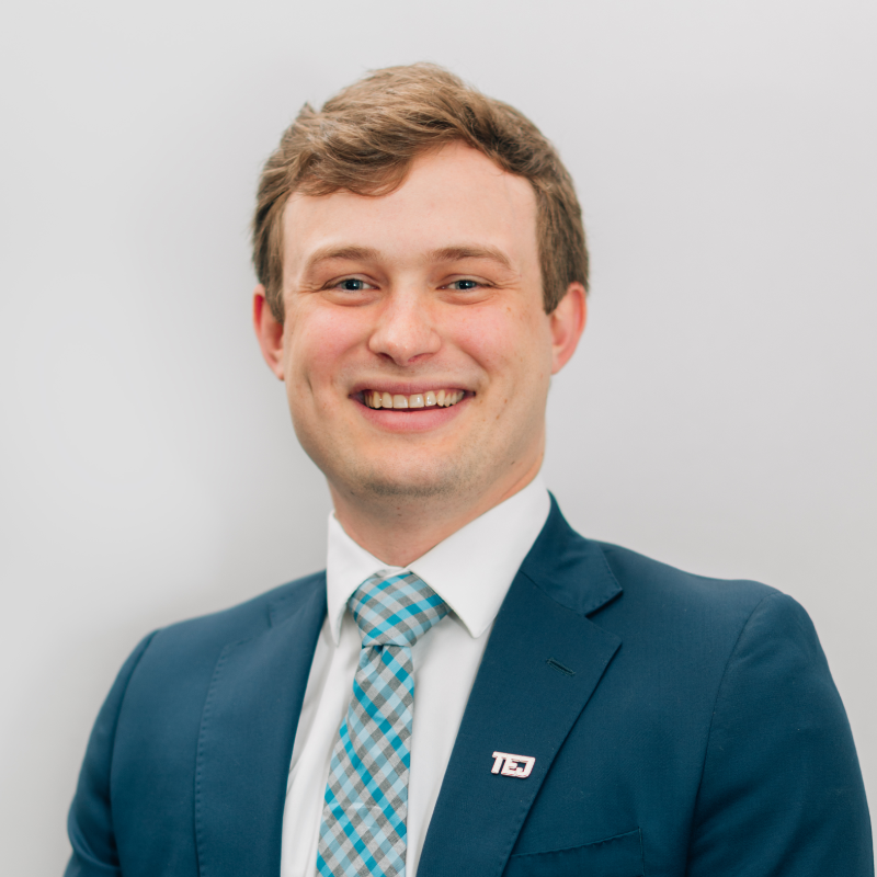 Smiling person in a blue suit and teal plaid tie against a light gray background