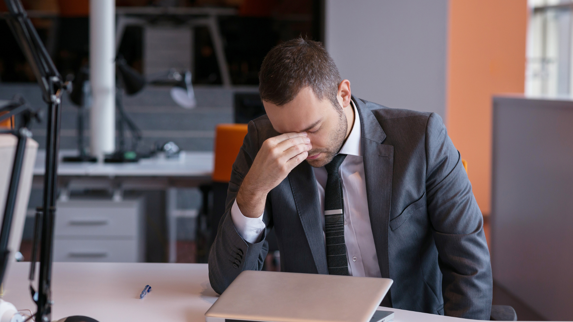 Stressed office worker sits at a desk, holding his head over an open laptop.