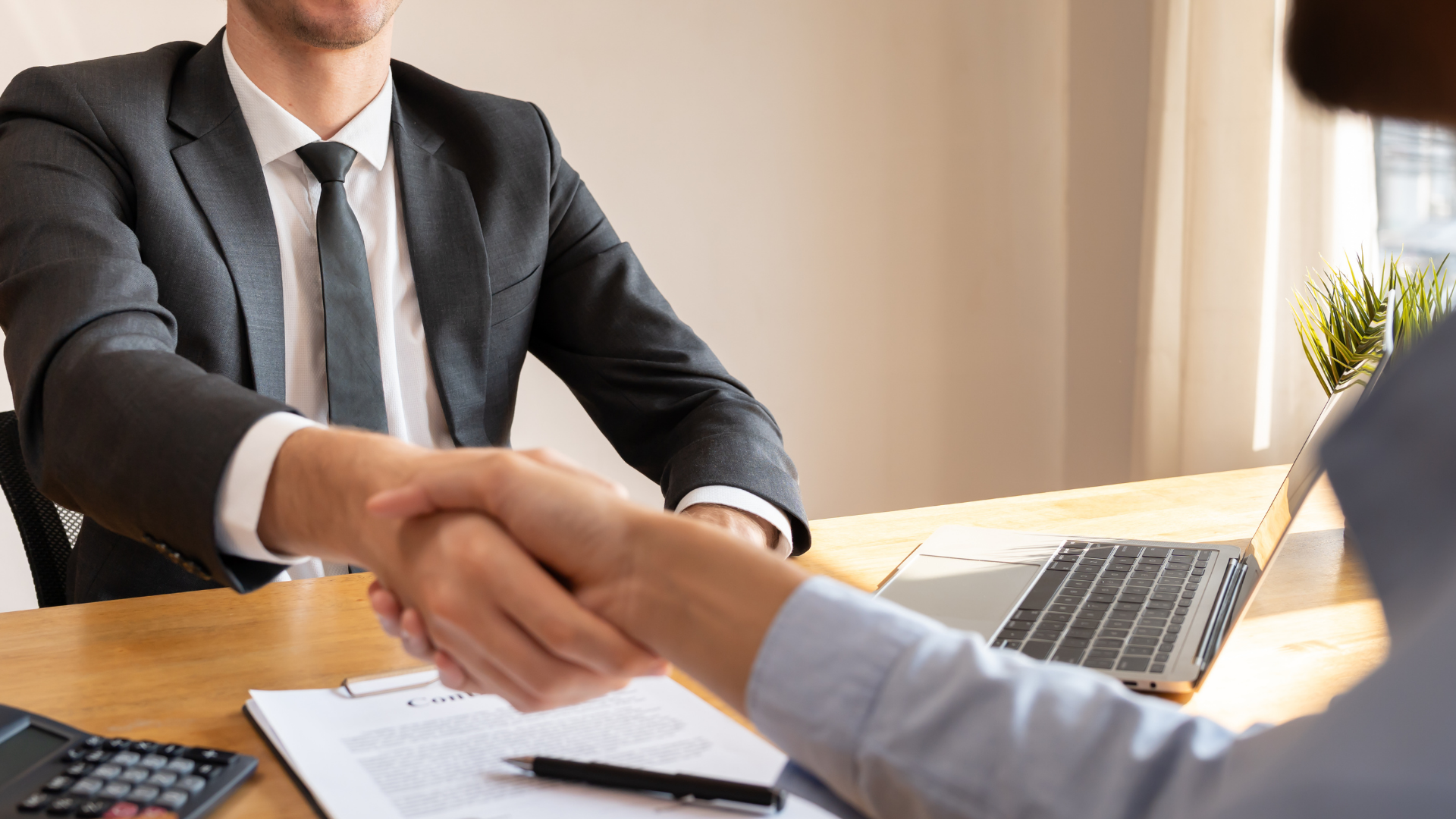 Two people shaking hands across a desk in an office, with a laptop and paperwork nearby