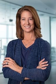 A woman in a blue sweater is standing with her arms crossed and smiling.