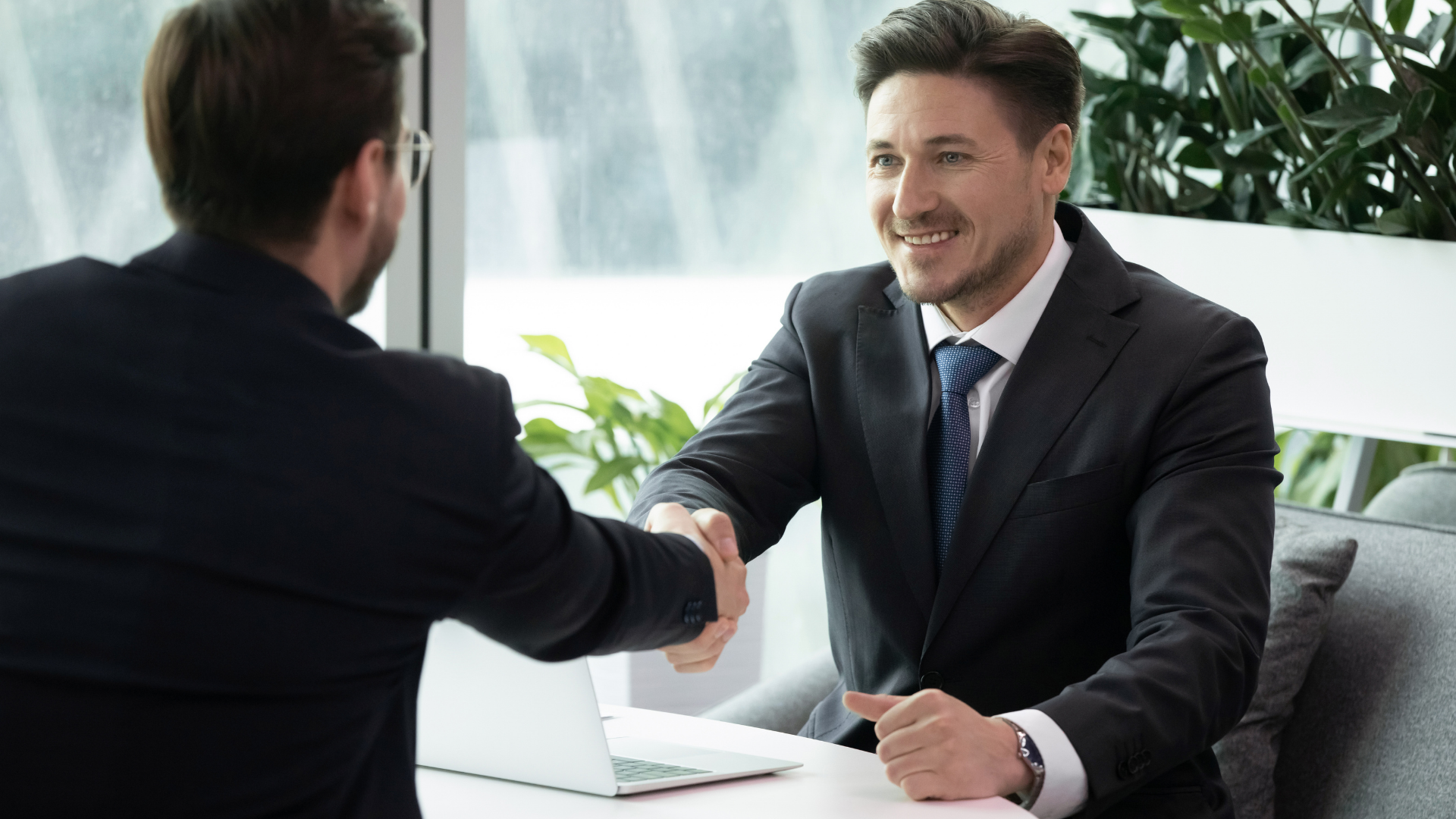 Two businessmen shaking hands in a bright office, with a laptop and potted plant nearby.