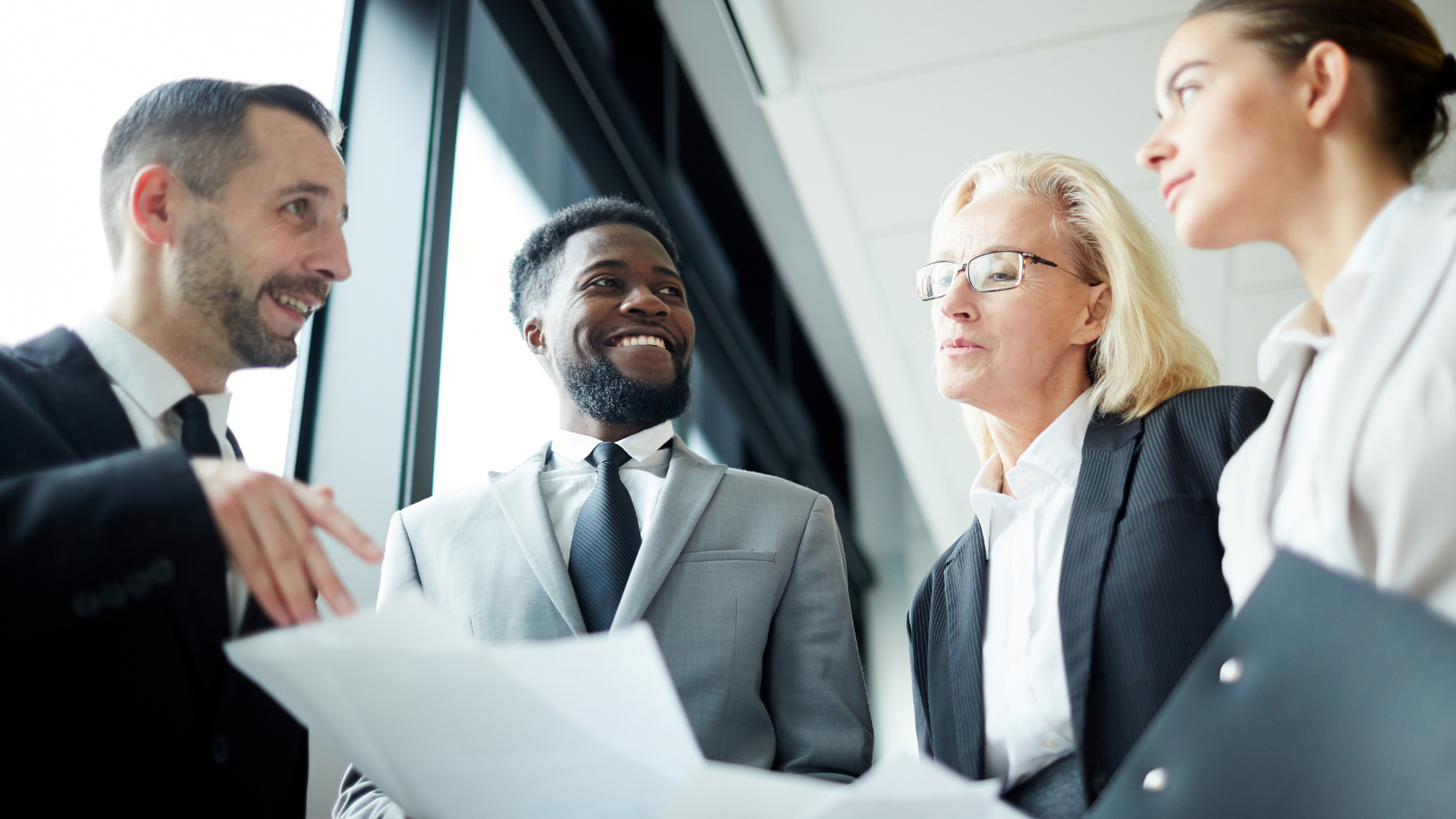 Four business professionals in suits talking in a bright office meeting