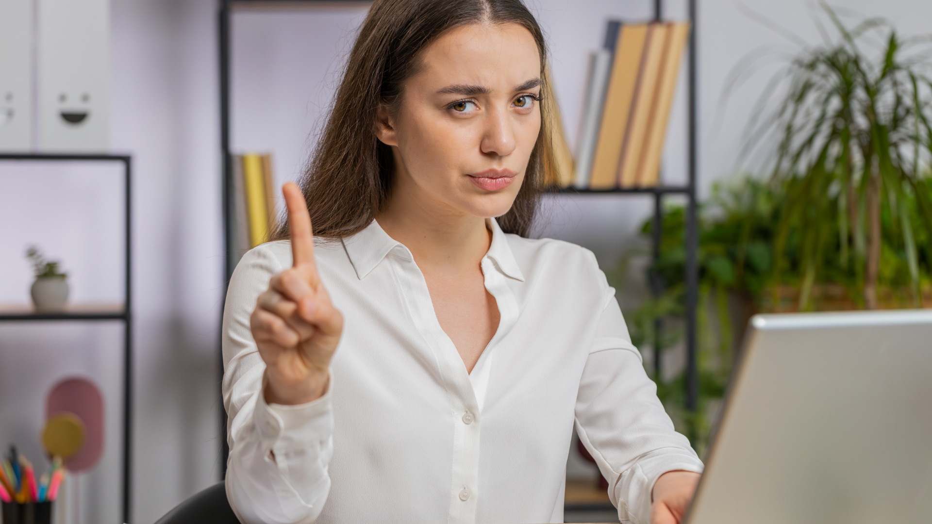 Woman in white shirt points index finger upward, appearing to make a request. Office setting with laptop and bookshelf visible.