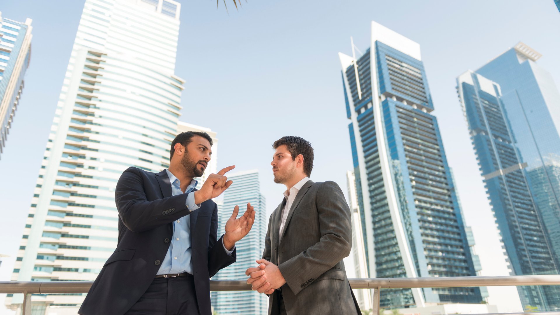 Two men in suits talking outdoors, pointing fingers. Skyscrapers in the background.