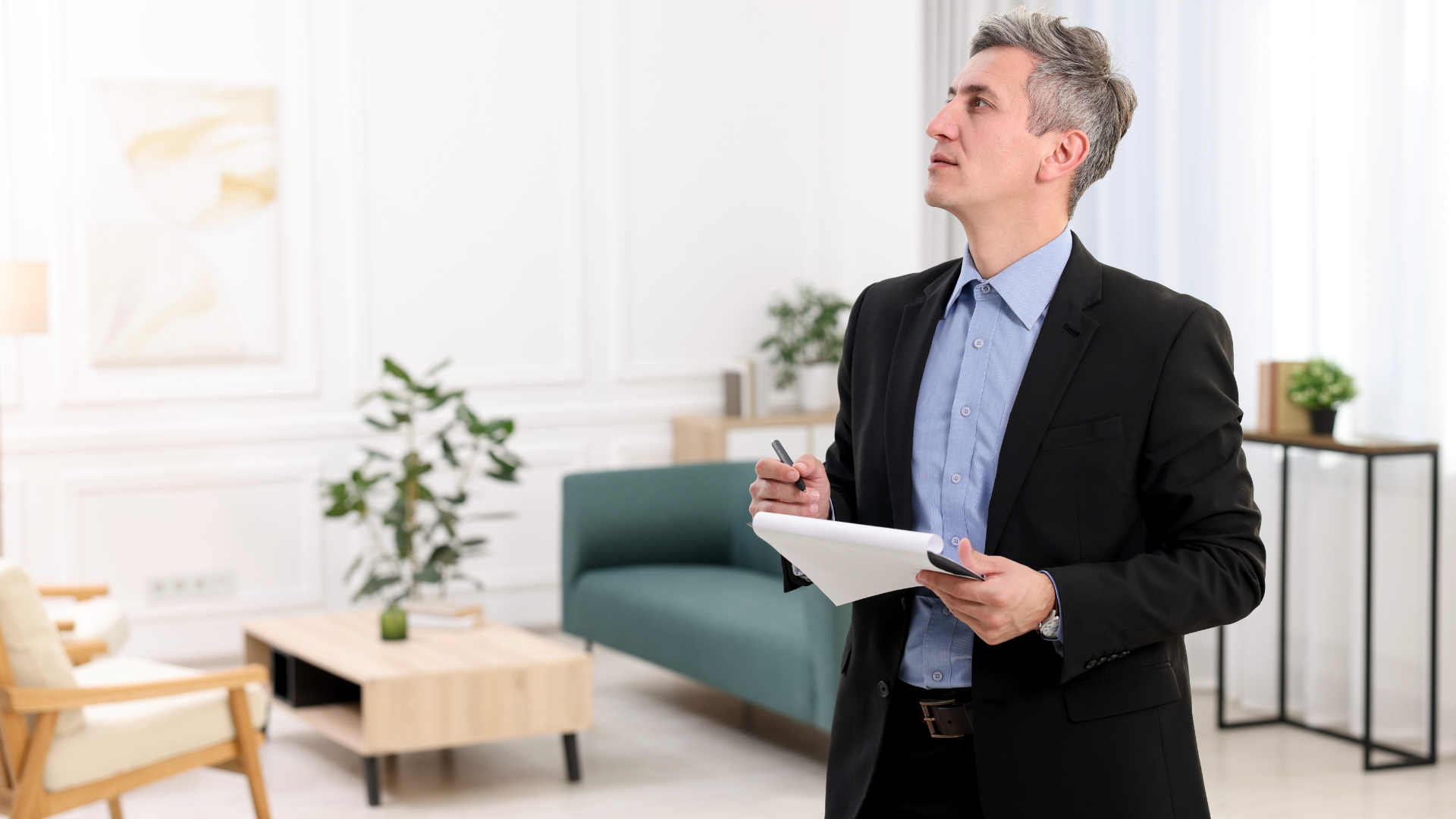 A man in a suit inspecting a room, holding a notepad, looking up.