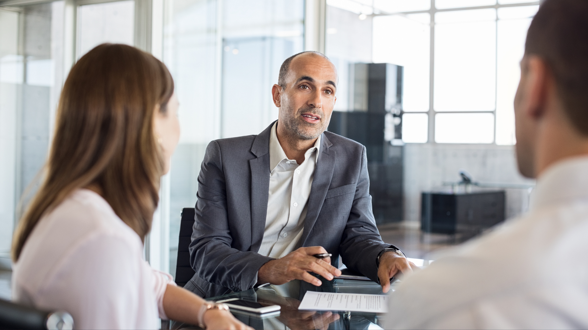 Business meeting: Man in blazer discusses documents with a couple at a table.