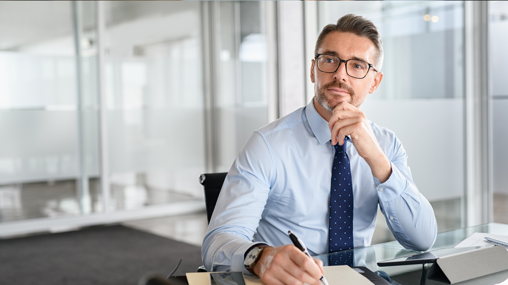 Man in blue shirt and tie sitting at a desk, holding a pen and looking thoughtfully at the camera.