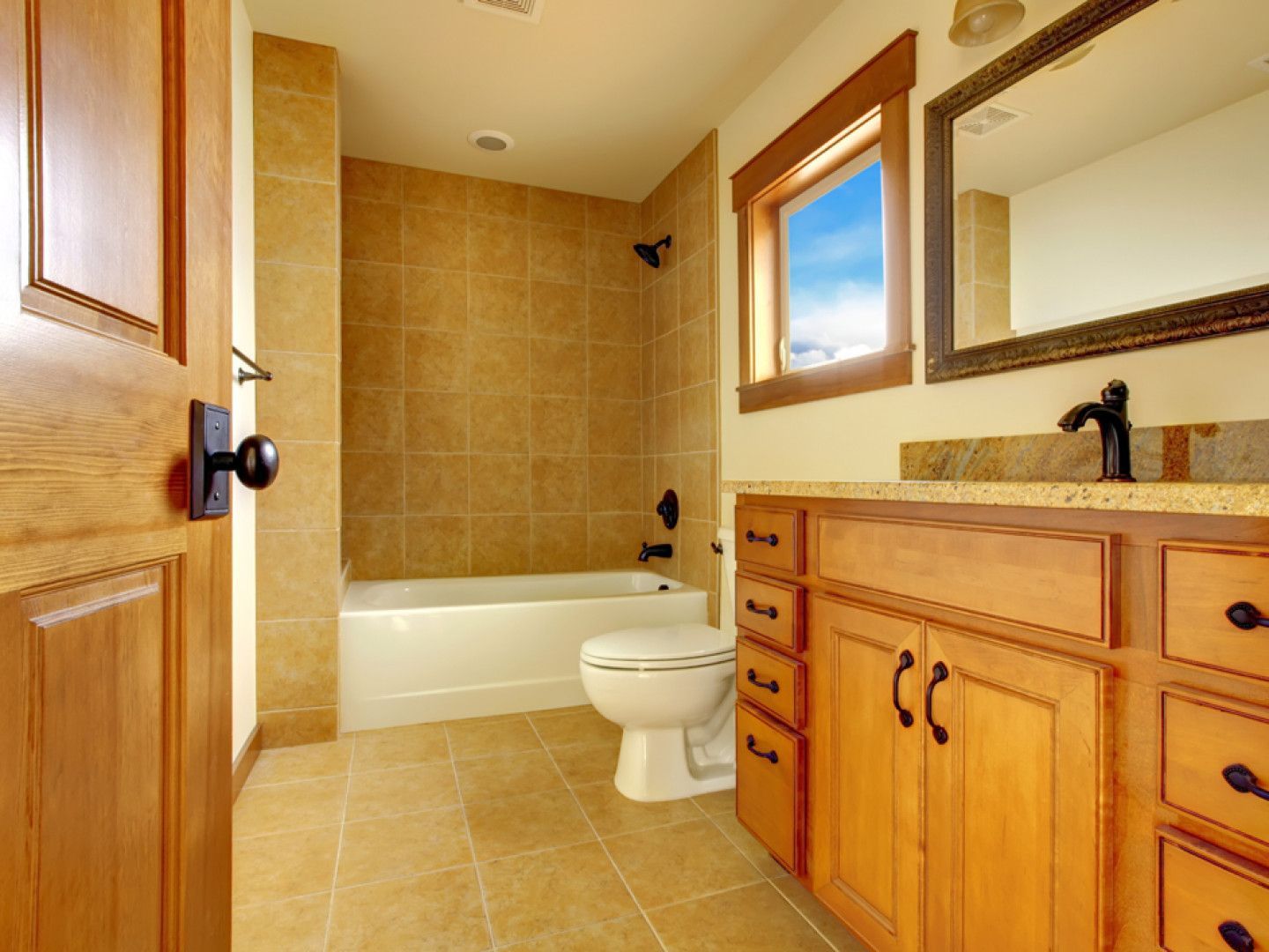Bathroom with tan tile, wooden vanity, and white tub and toilet; natural light.