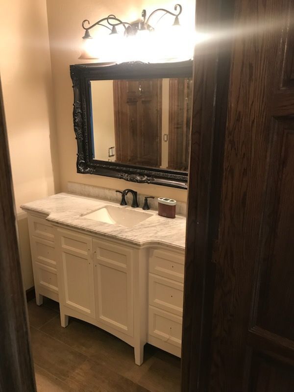 White bathroom vanity with marble top, black-framed mirror, and wood paneling on the wall.