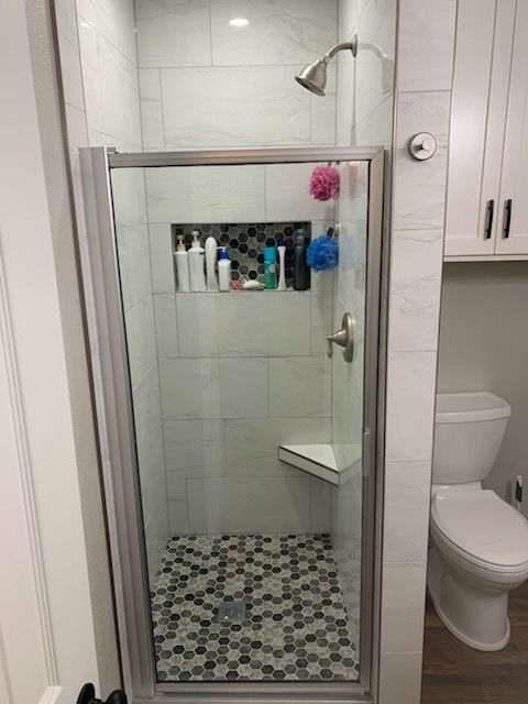 A tiled shower with a glass door, built-in shelf, and a corner seat. Toilet and cabinet nearby.