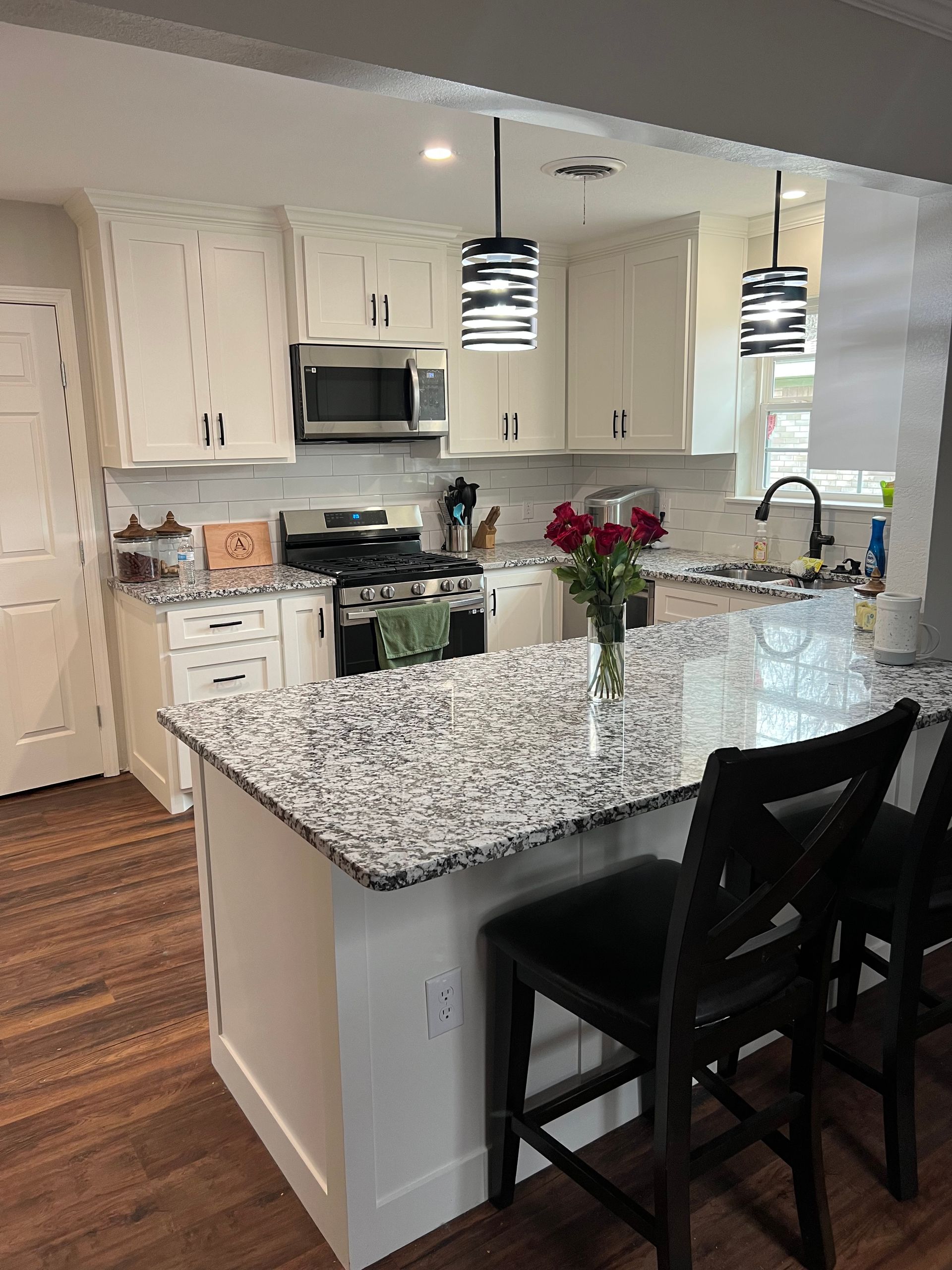 White kitchen with granite countertops and island, black bar stools, and pendant lights.
