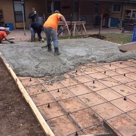 Workers pouring concrete for a patio, leveling with tools. Steel rebar grid visible in the foreground. Outdoors.