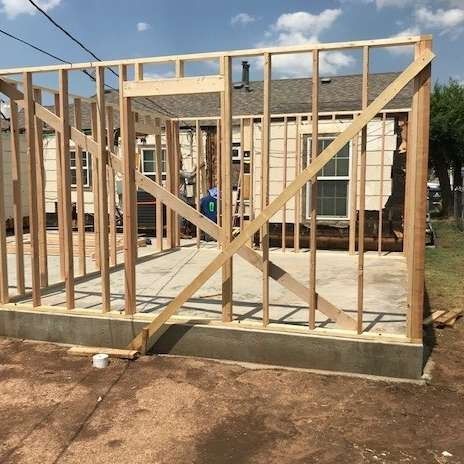 Wooden frame of a building under construction on a concrete foundation, with a house visible in the background.