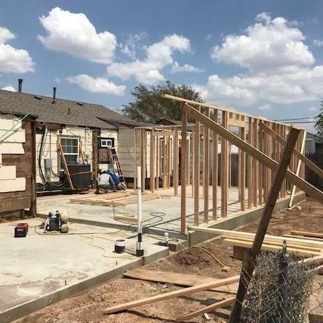Construction site: wooden frames rising from concrete foundation. Workers near partially demolished house on sunny day.