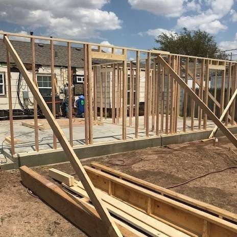 Construction of a wooden framed structure, likely a building, against a backdrop of another building and a blue sky.