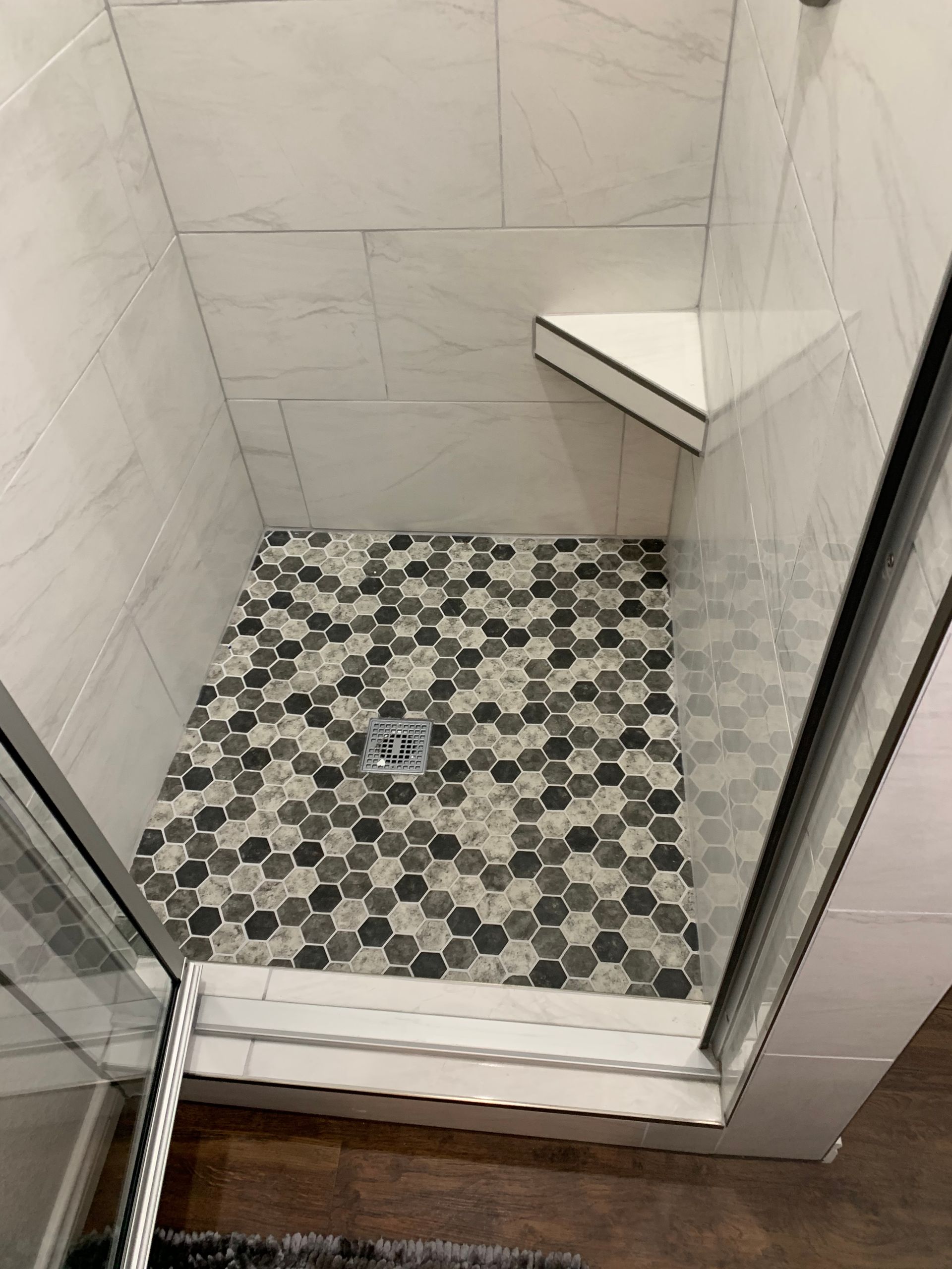 Shower with gray and black hexagon tile floor, white tiled walls, and corner seat.