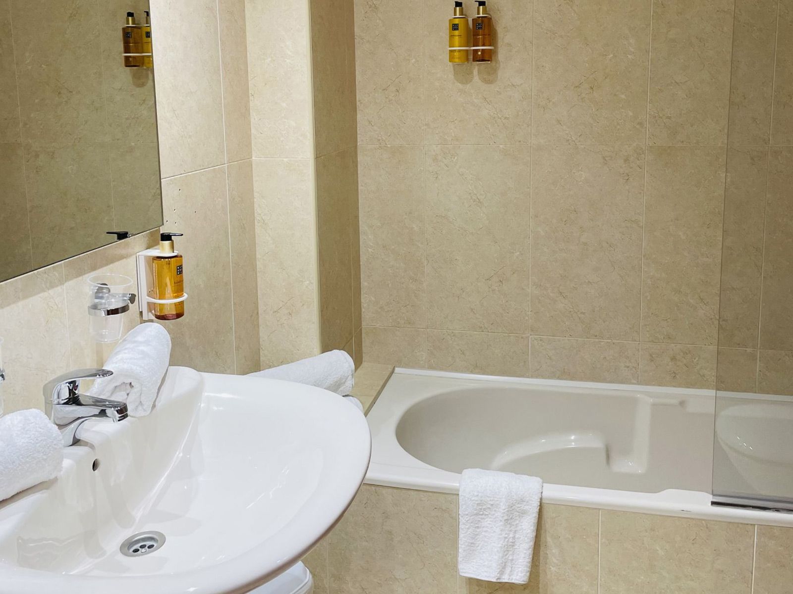 Baño con lavabo, encimera, bañera y paredes revestidas de azulejos beige; se ven toallas y artículos de aseo.