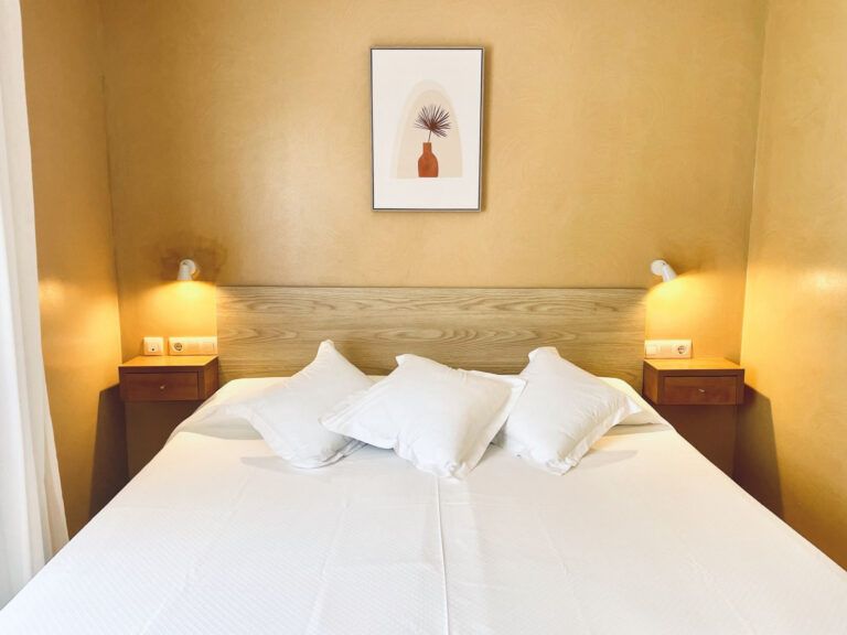 Dormitorio acogedor con una cama blanca bien hecha, lámparas de mesilla y una lámina enmarcada de flores sobre paredes beige