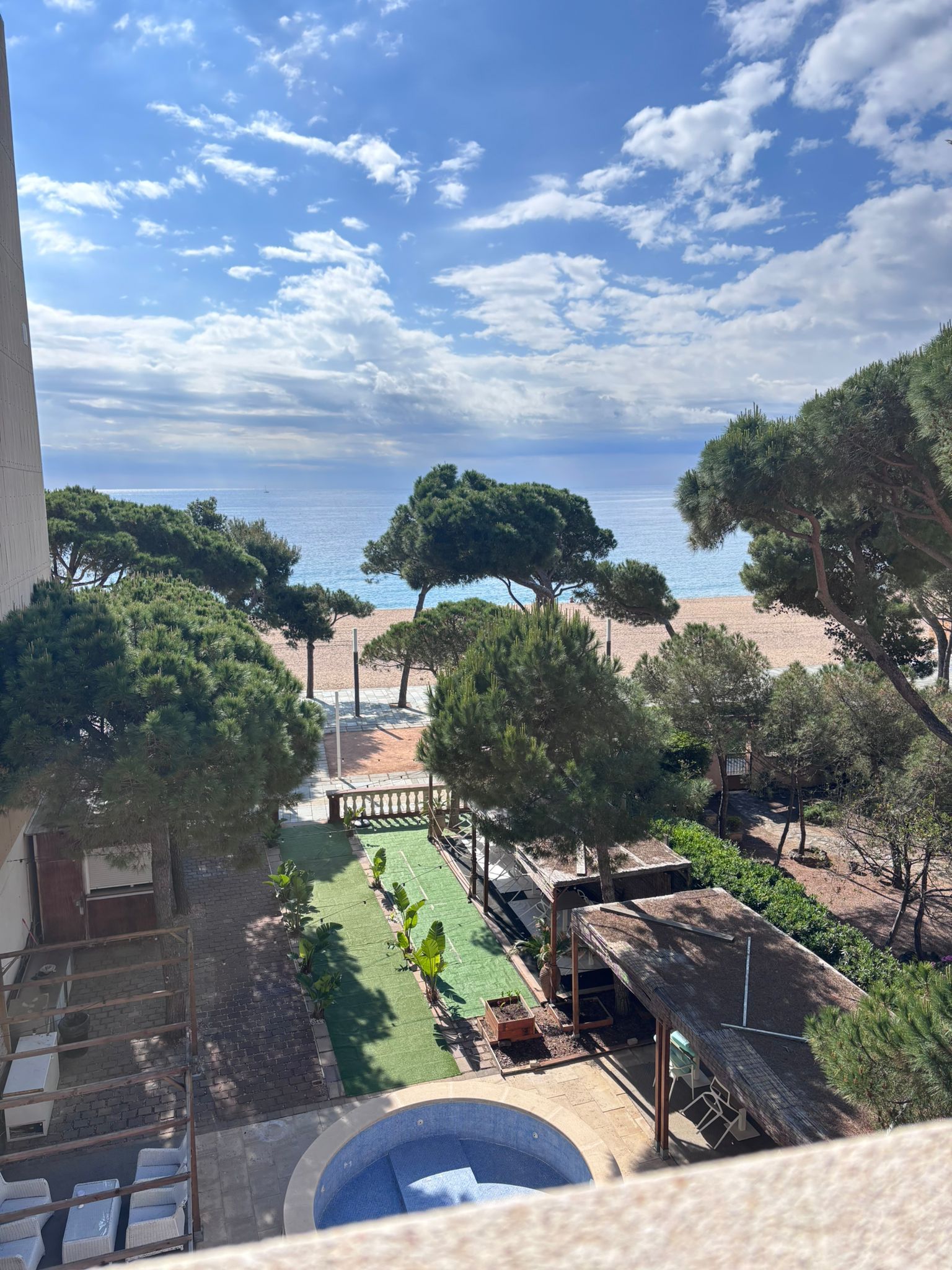 Vista desde un balcón de una piscina, palmeras y una playa de arena bajo un cielo azul despejado