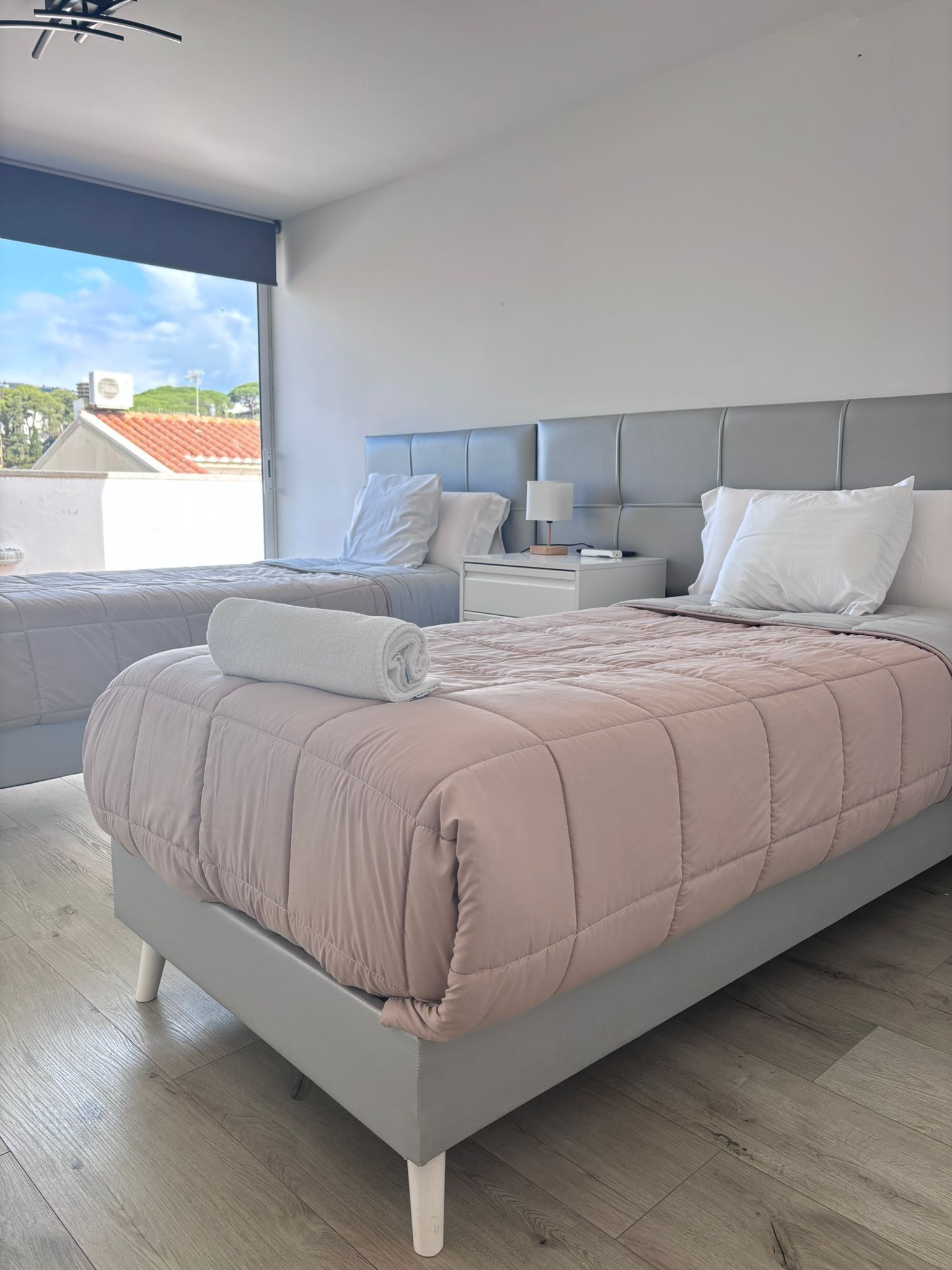 Dormitorio moderno y luminoso, con una colcha rosa, una cama tapizada en gris y una gran ventana por la que entra la luz.